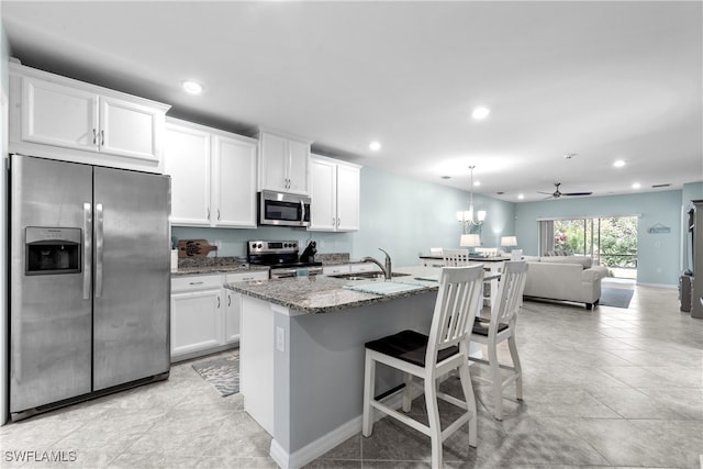 kitchen with white cabinetry, ceiling fan, stainless steel appliances, light stone counters, and a center island with sink