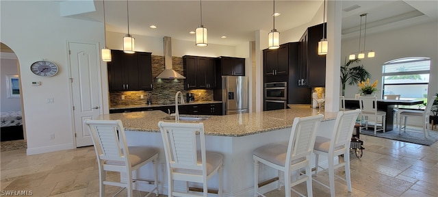 kitchen featuring stainless steel appliances, a breakfast bar area, wall chimney exhaust hood, backsplash, and light stone counters