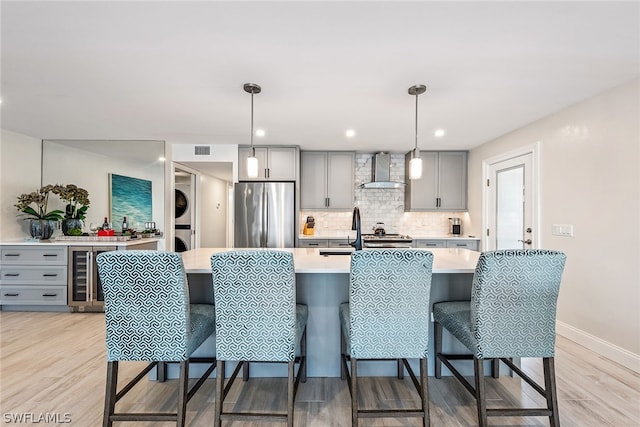 kitchen with stacked washer / drying machine, gray cabinets, stainless steel refrigerator, wine cooler, and wall chimney exhaust hood