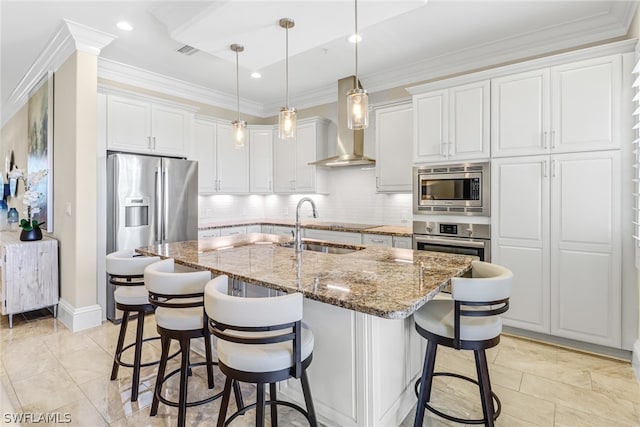 kitchen with white cabinets, appliances with stainless steel finishes, a kitchen island with sink, wall chimney exhaust hood, and sink