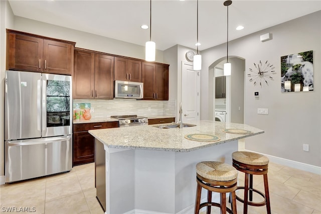 kitchen featuring a kitchen island with sink, appliances with stainless steel finishes, light stone counters, sink, and decorative light fixtures