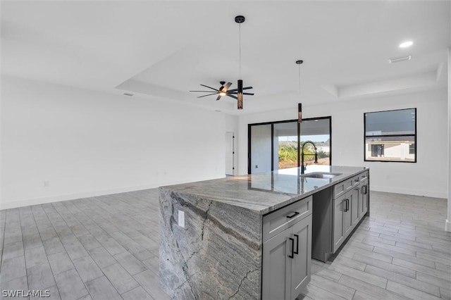 kitchen with light stone counters, a tray ceiling, an island with sink, gray cabinetry, and a sink