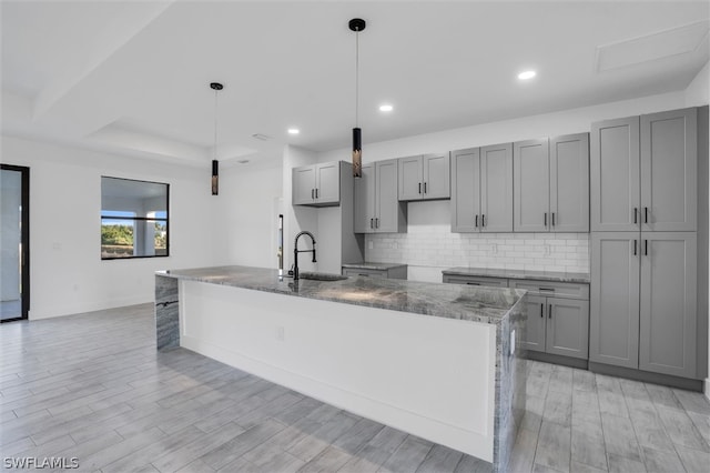 kitchen with dark stone counters, a kitchen island with sink, sink, pendant lighting, and light hardwood / wood-style flooring