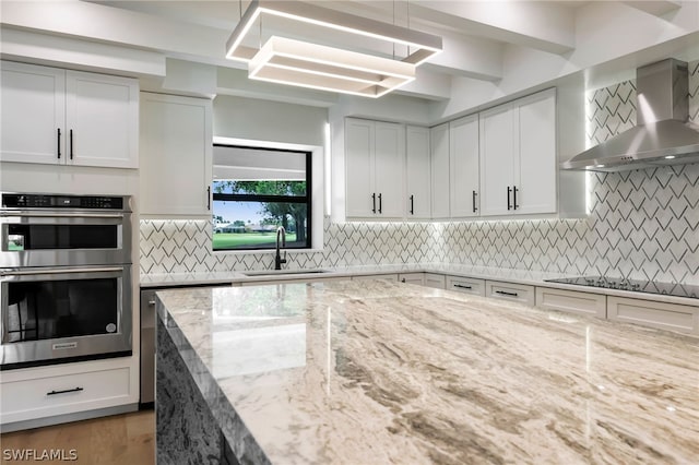 kitchen with light stone counters, black electric cooktop, stainless steel double oven, wall chimney range hood, and a sink