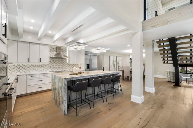 kitchen featuring light wood finished floors, decorative backsplash, stainless steel built in fridge, wall chimney range hood, and beamed ceiling