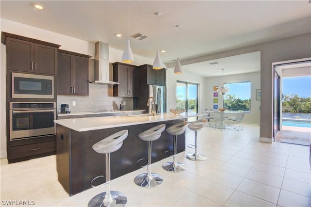 kitchen featuring a kitchen island with sink, appliances with stainless steel finishes, a breakfast bar, wall chimney range hood, and decorative light fixtures