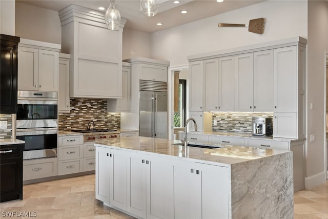 kitchen with pendant lighting, stainless steel appliances, sink, a kitchen island with sink, and light stone counters