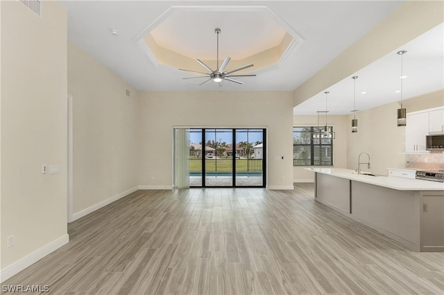 unfurnished living room with light wood-type flooring, a tray ceiling, sink, and ceiling fan with notable chandelier