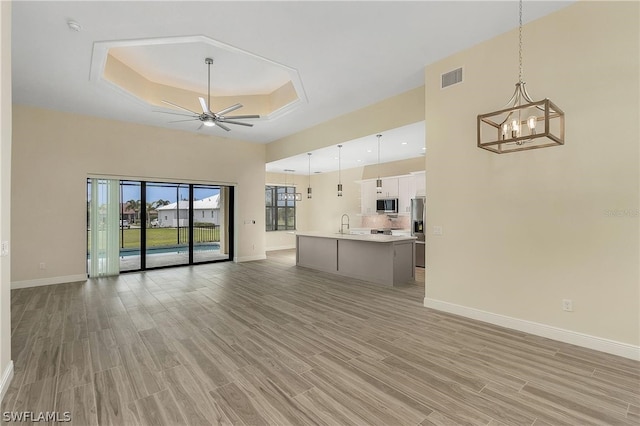 unfurnished living room featuring light hardwood / wood-style floors, ceiling fan with notable chandelier, sink, and a tray ceiling