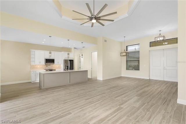 kitchen with ceiling fan with notable chandelier, appliances with stainless steel finishes, white cabinetry, a tray ceiling, and a kitchen island with sink