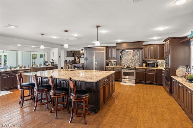 kitchen featuring pendant lighting, an island with sink, a breakfast bar area, built in appliances, and light stone countertops