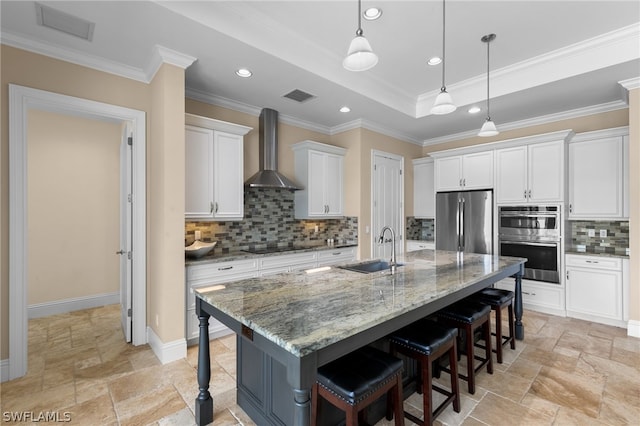 kitchen with stainless steel appliances, wall chimney range hood, a kitchen bar, a kitchen island with sink, and white cabinets