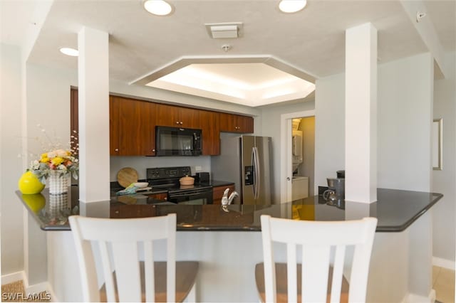 kitchen featuring a kitchen breakfast bar, black appliances, stacked washer / drying machine, a tray ceiling, and kitchen peninsula