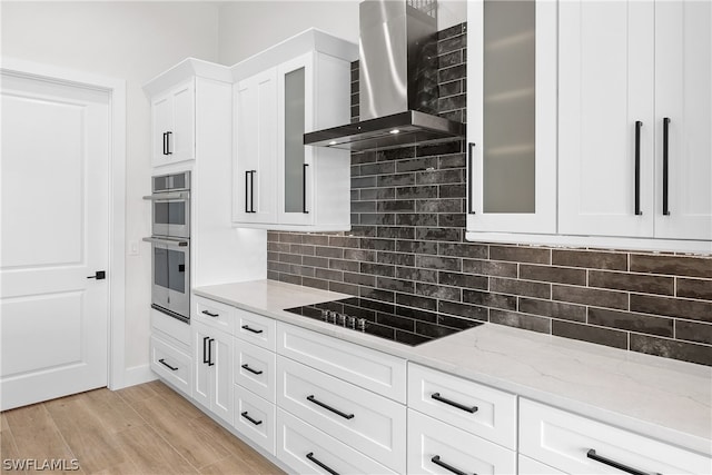 kitchen featuring double oven, wall chimney range hood, tasteful backsplash, black electric cooktop, and light hardwood / wood-style floors