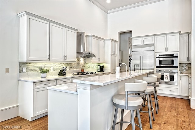 kitchen featuring light wood-type flooring, wall chimney range hood, a breakfast bar, stainless steel appliances, and a center island with sink