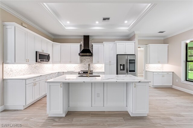 kitchen featuring wall chimney range hood, backsplash, a center island with sink, light wood-type flooring, and appliances with stainless steel finishes