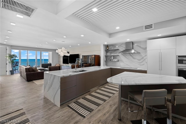 kitchen featuring wall chimney range hood, light stone counters, sink, white cabinetry, and light hardwood / wood-style flooring