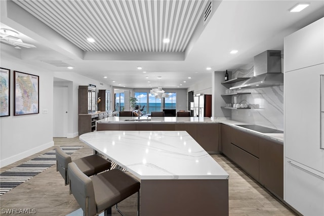 kitchen featuring wall chimney range hood, a center island, black electric stovetop, a kitchen bar, and light hardwood / wood-style flooring