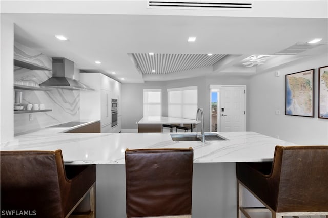 kitchen featuring a raised ceiling, a kitchen bar, sink, light stone countertops, and wall chimney exhaust hood