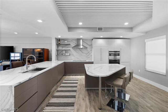 kitchen with sink, wall chimney exhaust hood, wood-type flooring, stainless steel double oven, and light stone counters