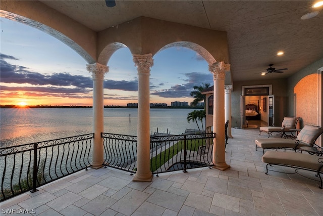 patio terrace at dusk featuring ceiling fan, a water view, and a balcony