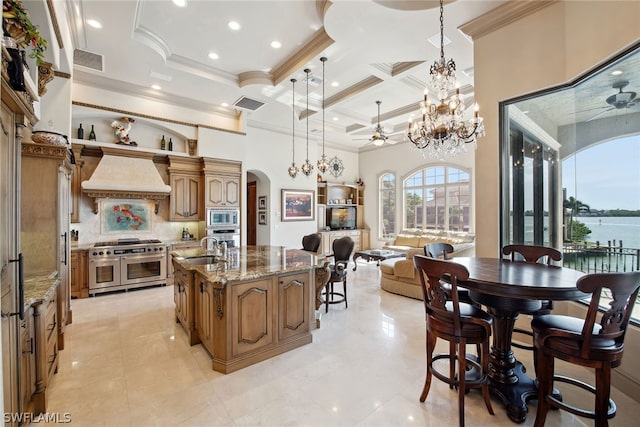 kitchen featuring custom exhaust hood, ceiling fan with notable chandelier, stainless steel appliances, dark stone countertops, and a kitchen island with sink