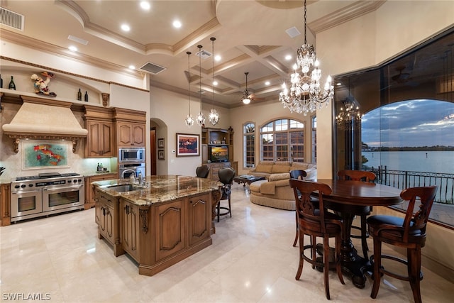 kitchen featuring custom exhaust hood, ceiling fan with notable chandelier, a water view, appliances with stainless steel finishes, and coffered ceiling