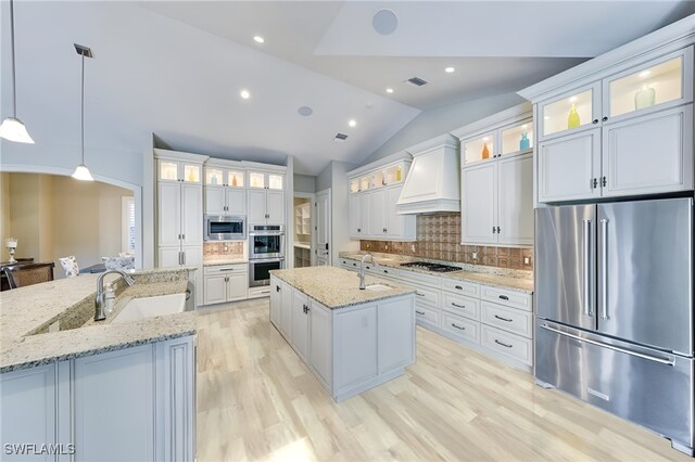 kitchen featuring pendant lighting, custom exhaust hood, vaulted ceiling, an island with sink, and appliances with stainless steel finishes