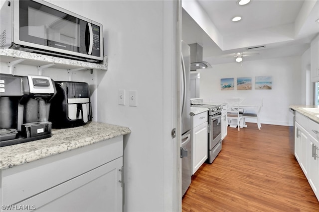 kitchen featuring ceiling fan, wall chimney exhaust hood, light hardwood / wood-style flooring, white cabinetry, and stainless steel appliances