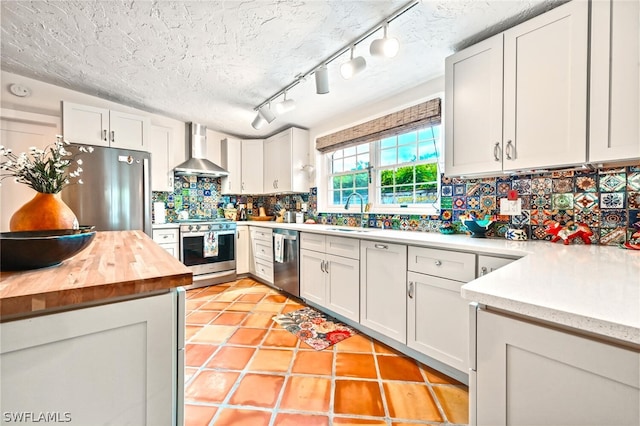 kitchen featuring stainless steel appliances, white cabinets, a sink, butcher block countertops, and wall chimney exhaust hood