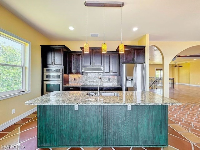 kitchen with pendant lighting, light stone countertops, stainless steel appliances, a center island with sink, and tasteful backsplash