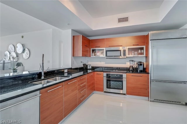 kitchen featuring appliances with stainless steel finishes, a tray ceiling, dark stone countertops, sink, and light tile floors