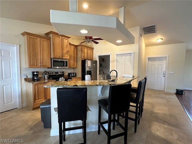 kitchen with a breakfast bar, stainless steel appliances, light stone counters, ceiling fan, and an island with sink