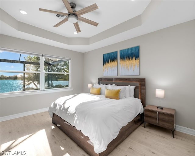 bedroom featuring light wood-type flooring, a water view, a raised ceiling, and baseboards