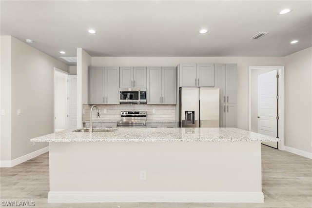 kitchen featuring appliances with stainless steel finishes, light stone counters, sink, a center island with sink, and light hardwood / wood-style flooring