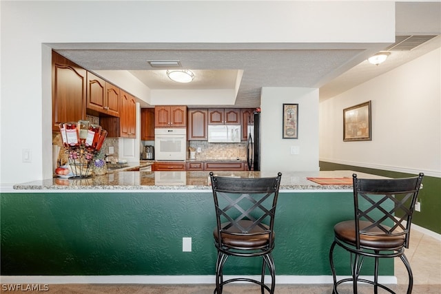 kitchen featuring light stone counters, light tile floors, white appliances, backsplash, and kitchen peninsula