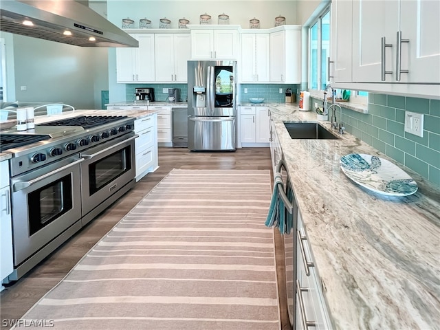 kitchen featuring white cabinets, wall chimney range hood, stainless steel appliances, sink, and light stone counters