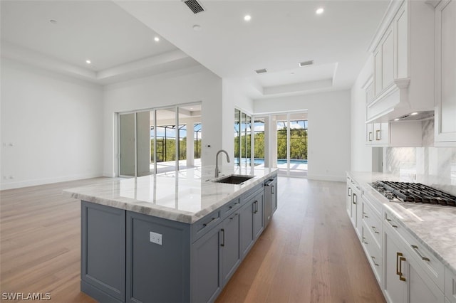 kitchen featuring sink, white cabinetry, light stone counters, a center island with sink, and a raised ceiling