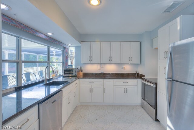 kitchen with stainless steel appliances, dark stone counters, white cabinets, sink, and light tile floors