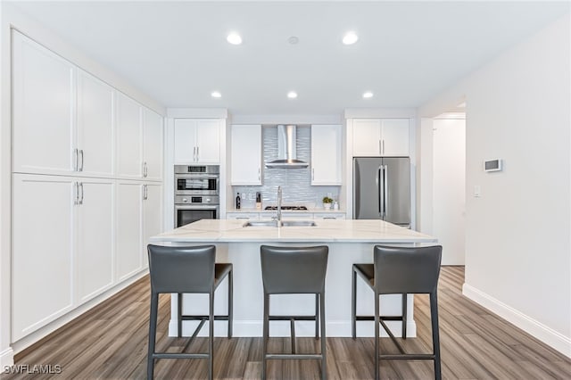 kitchen with appliances with stainless steel finishes, a center island with sink, white cabinetry, and wall chimney range hood