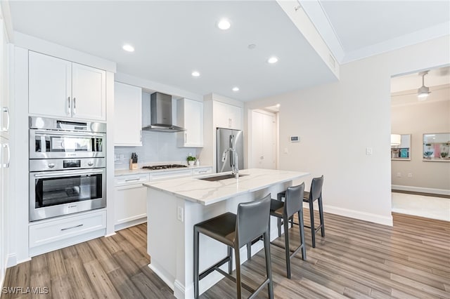 kitchen with appliances with stainless steel finishes, sink, wall chimney range hood, white cabinets, and an island with sink