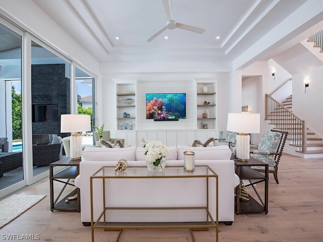 living room featuring built in shelves, ceiling fan, a raised ceiling, and light wood-type flooring