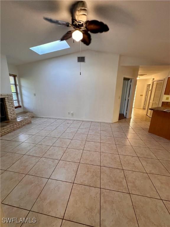 unfurnished living room with light tile patterned floors, ceiling fan, vaulted ceiling with skylight, and a brick fireplace