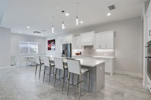 kitchen featuring custom range hood, backsplash, hanging light fixtures, a kitchen island with sink, and white cabinets