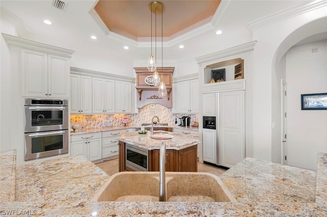 kitchen with built in appliances, tasteful backsplash, an island with sink, light stone counters, and a raised ceiling