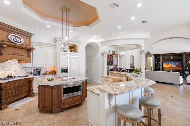 kitchen with white cabinets, backsplash, built in appliances, an island with sink, and light stone countertops