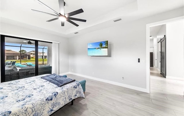 bedroom featuring ceiling fan, a barn door, light hardwood / wood-style floors, access to exterior, and a tray ceiling