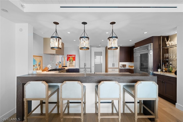 kitchen with kitchen peninsula, a kitchen bar, stainless steel built in fridge, light hardwood / wood-style floors, and dark brown cabinetry