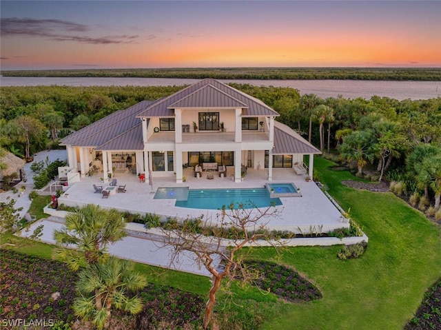 back house at dusk with a balcony, a patio, a pool with hot tub, a water view, and a yard