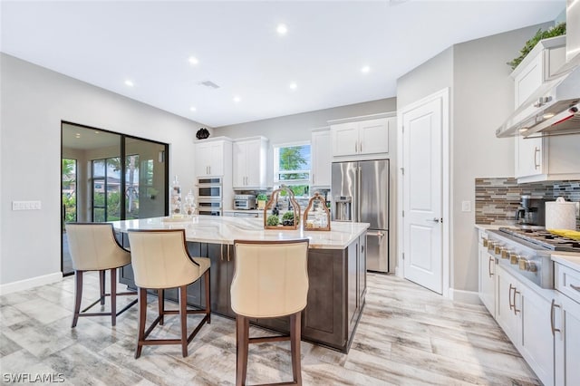 kitchen featuring wall chimney range hood, stainless steel appliances, white cabinets, and an island with sink